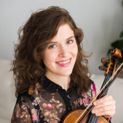 A white woman with brown hair holding a violin and smiling.