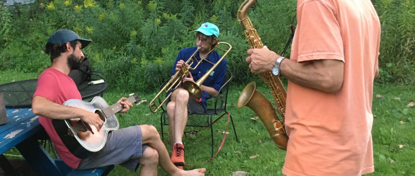 Three male figures jam in a backyard, playing guitar, saxophone, and trombone.