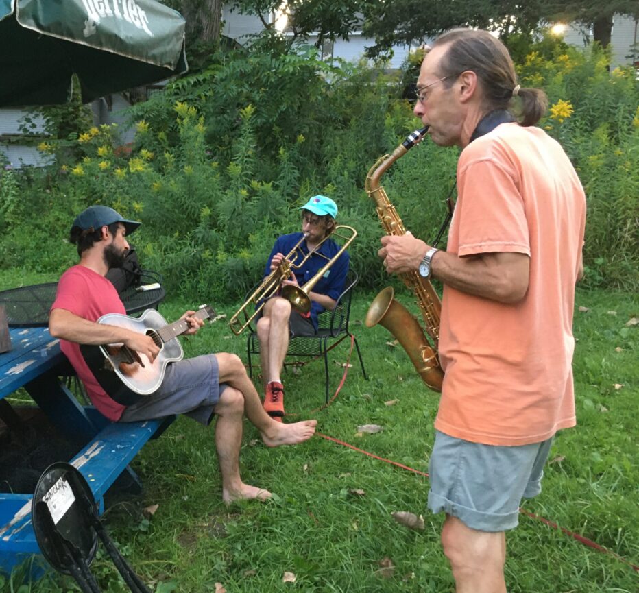 Three male figures jam in a backyard, playing guitar, saxophone, and trombone.